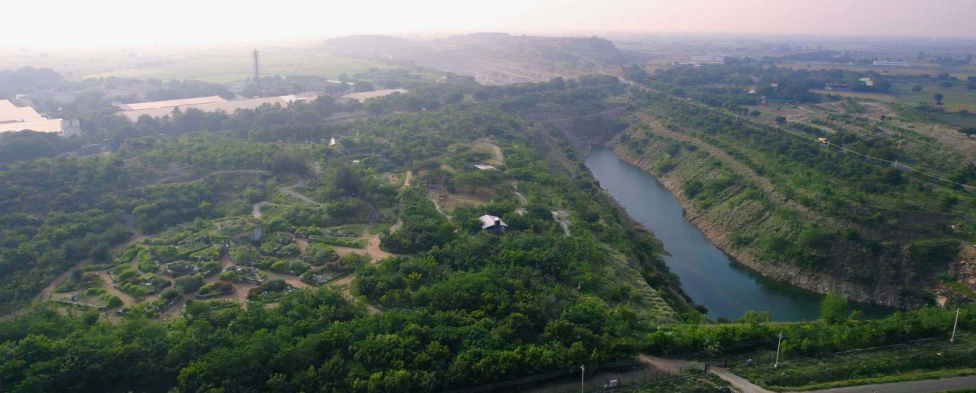 Aerial view of a restored quarry landscape: native forest and garden beds beside a water-filled pit, with farmland beyond