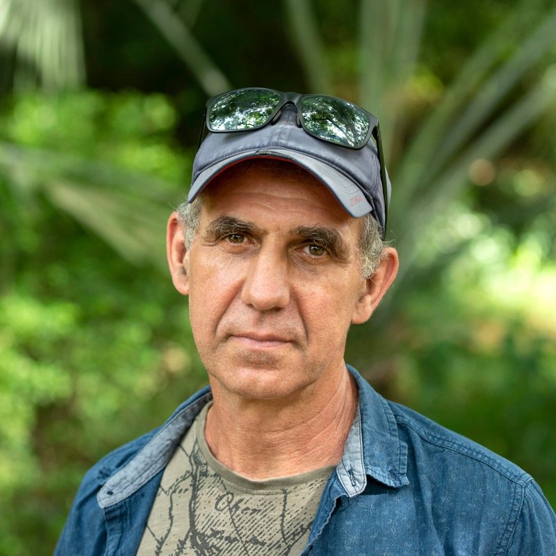 Portrait of Paul Blanchflower in a cap and denim shirt, with forest foliage behind